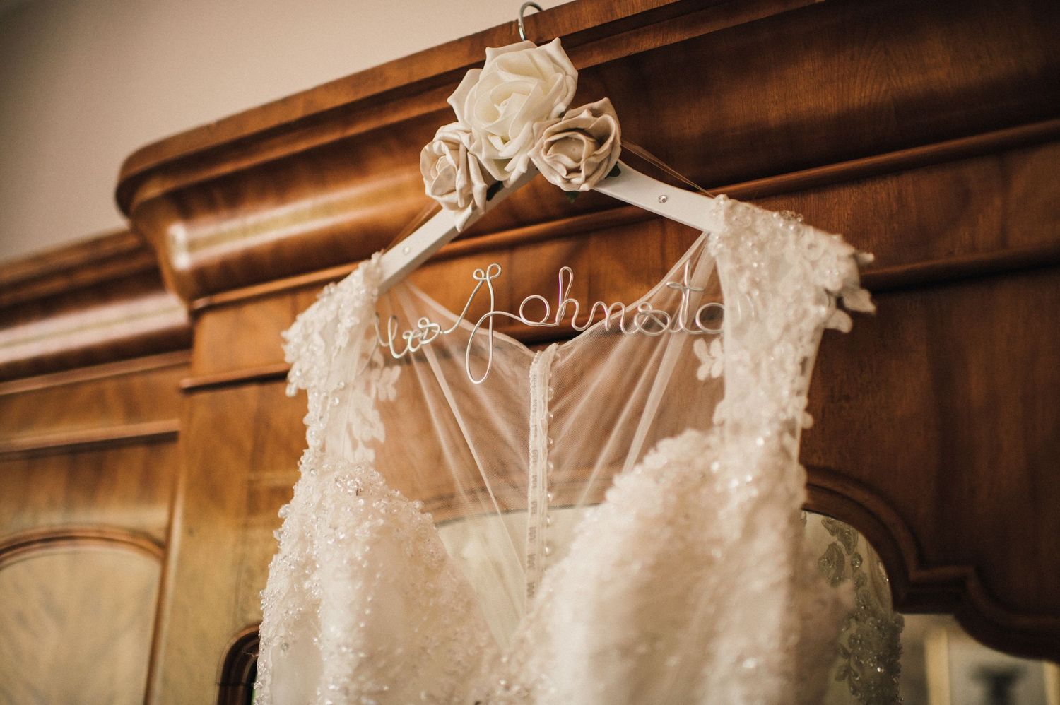 A detailed lace wedding dress hangs on a decorative hanger against wooden furniture.