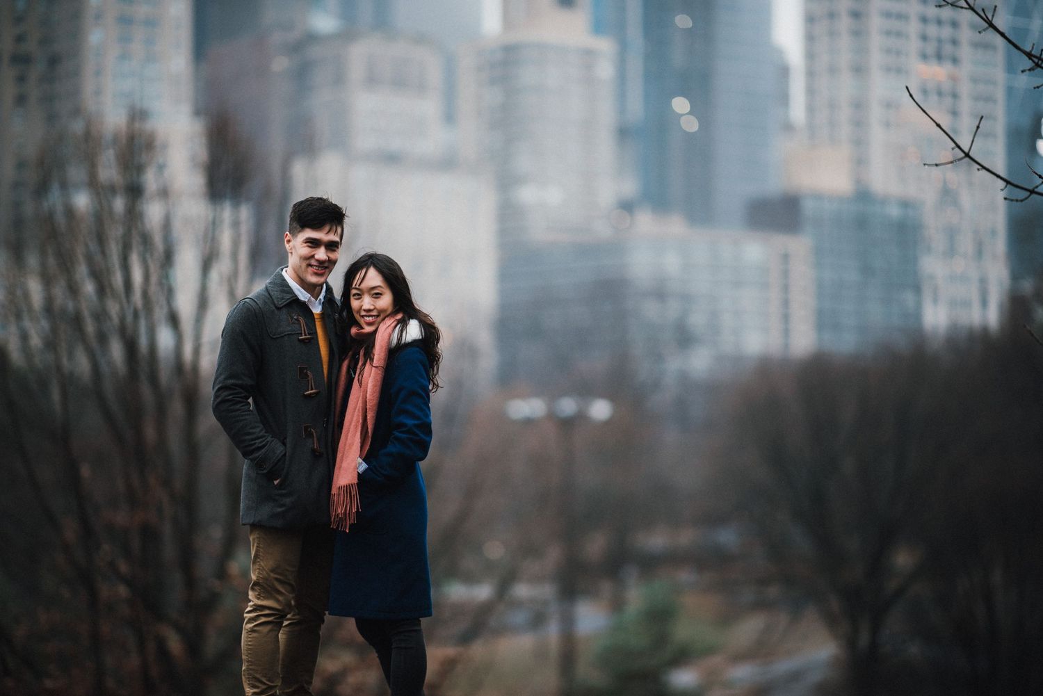 A couple poses outdoors with a blurred urban skyline backdrop.