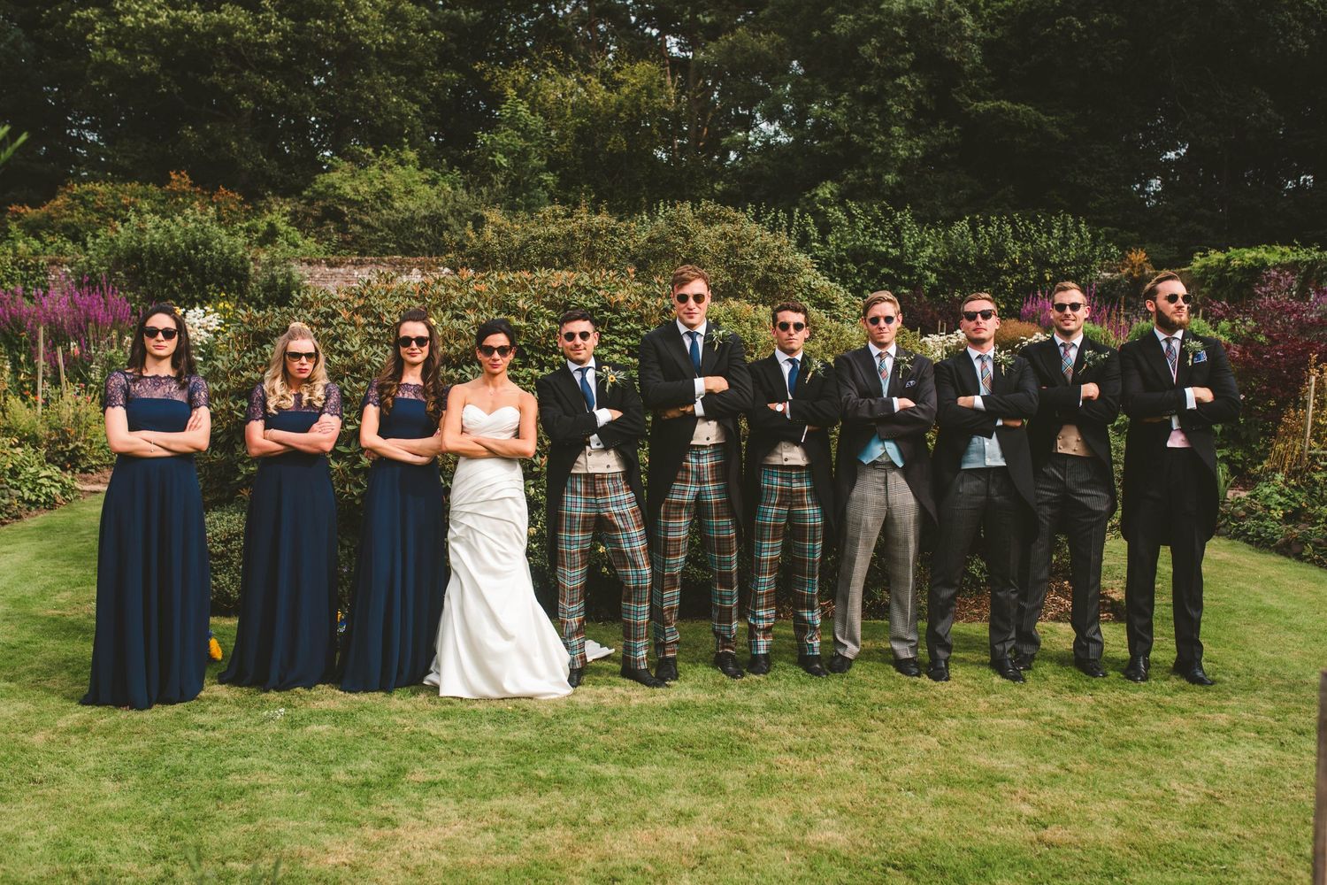 Wedding party poses outdoors wearing navy dresses and traditional Scottish kilts in a garden setting.