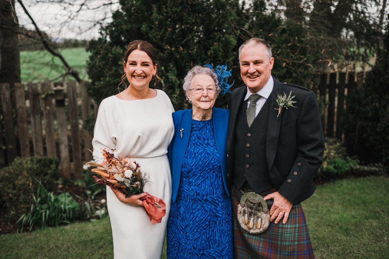 Three people pose for formal wedding photo in Scottish attire against garden backdrop.