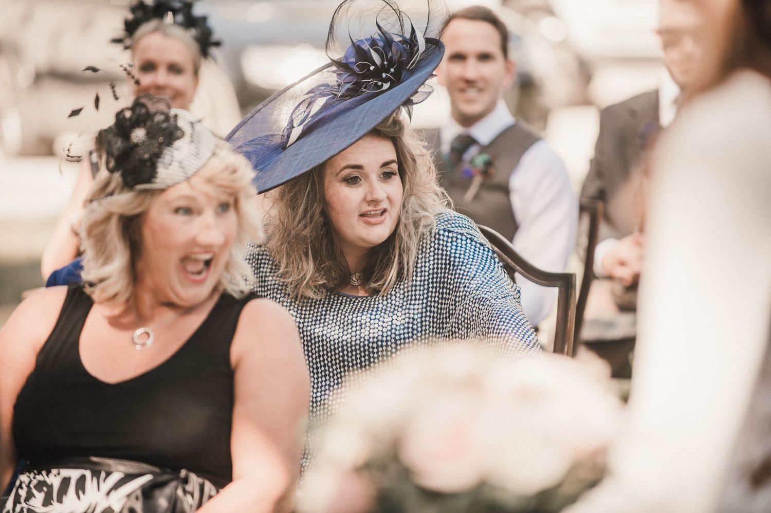 Wedding guests in formal attire and fancy hats enjoy the outdoor celebration.