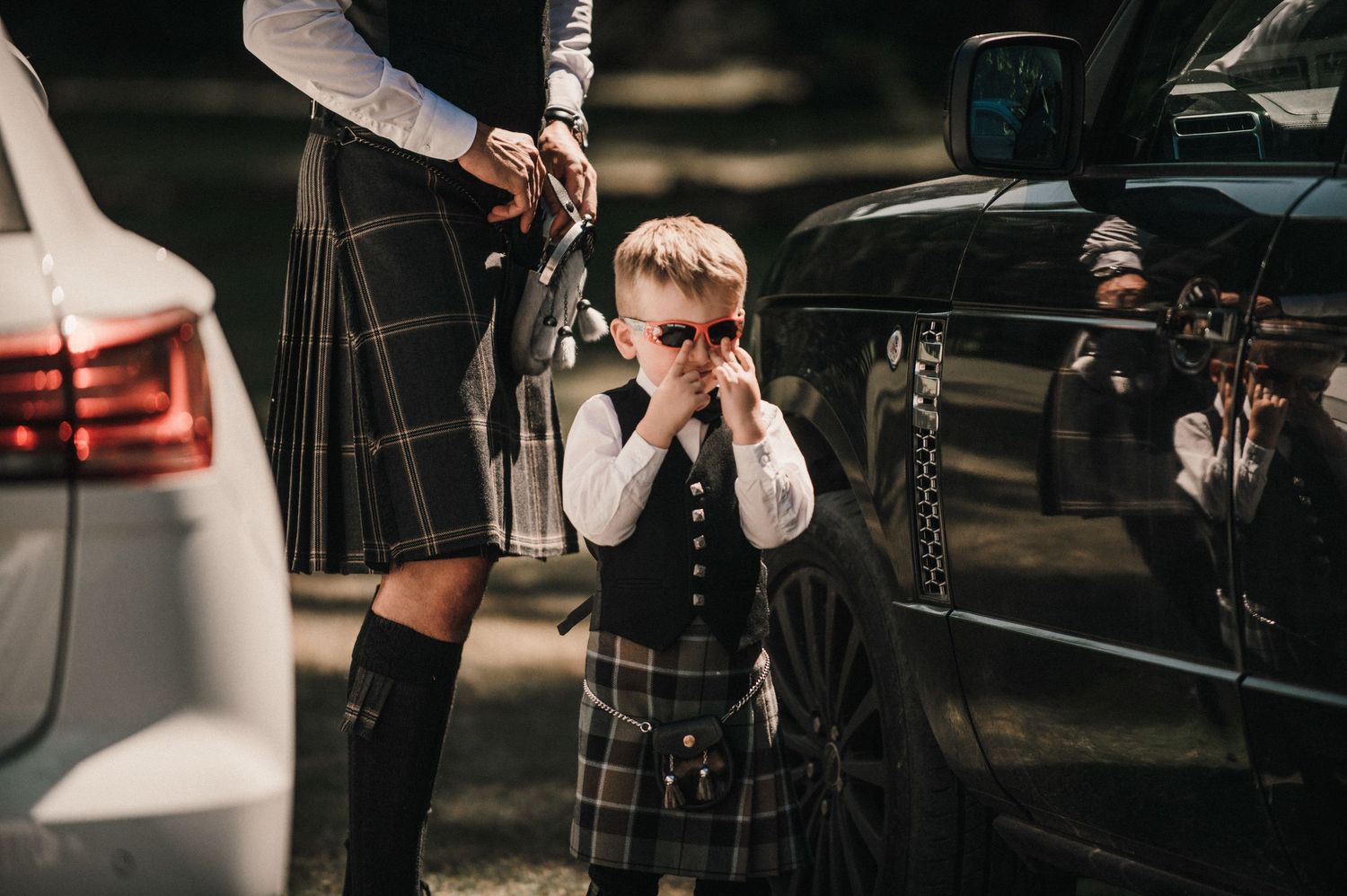 Small child in traditional Scottish kilt covering face while standing by cars.