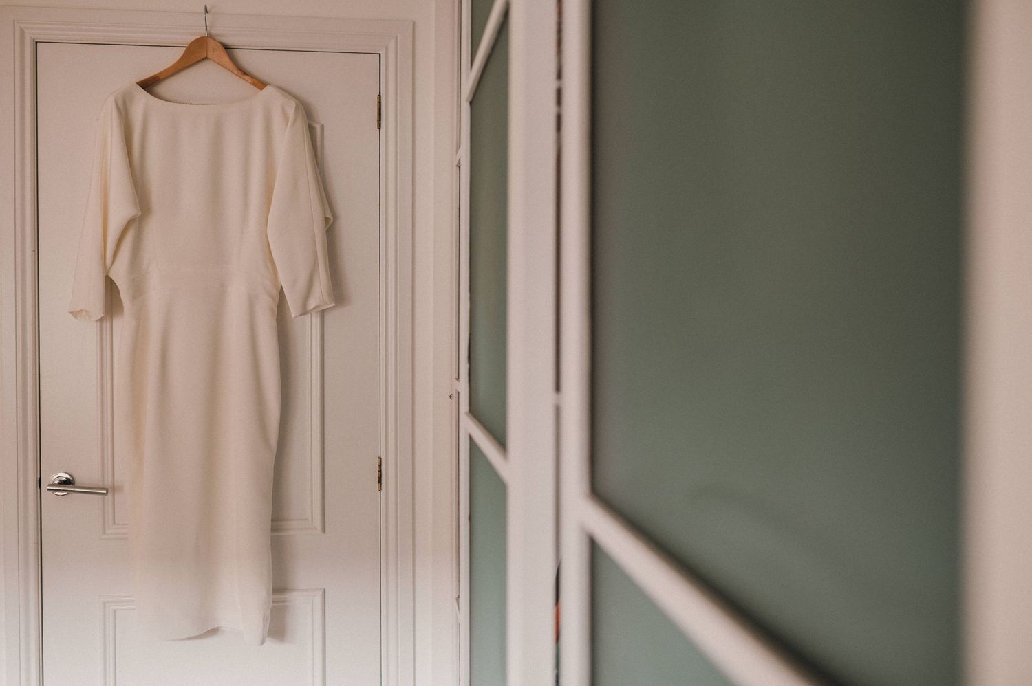 White elegant dress hanging on a wooden hanger against a white door.