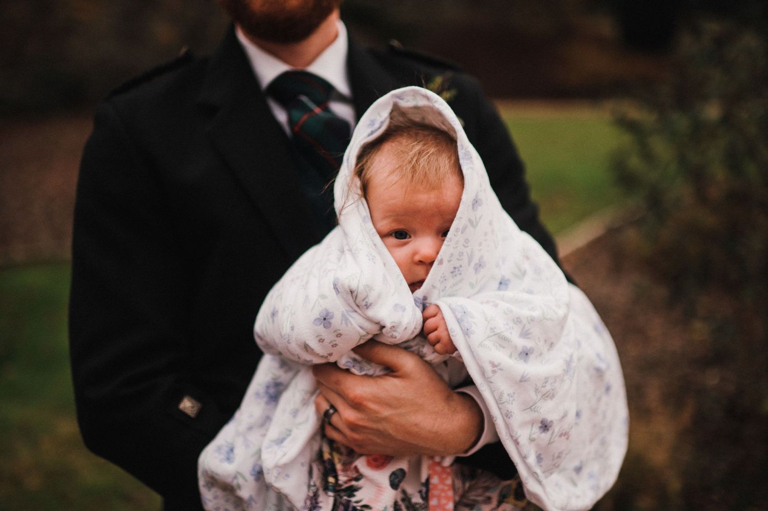 A small baby wrapped in white lace is held against formal wedding attire.