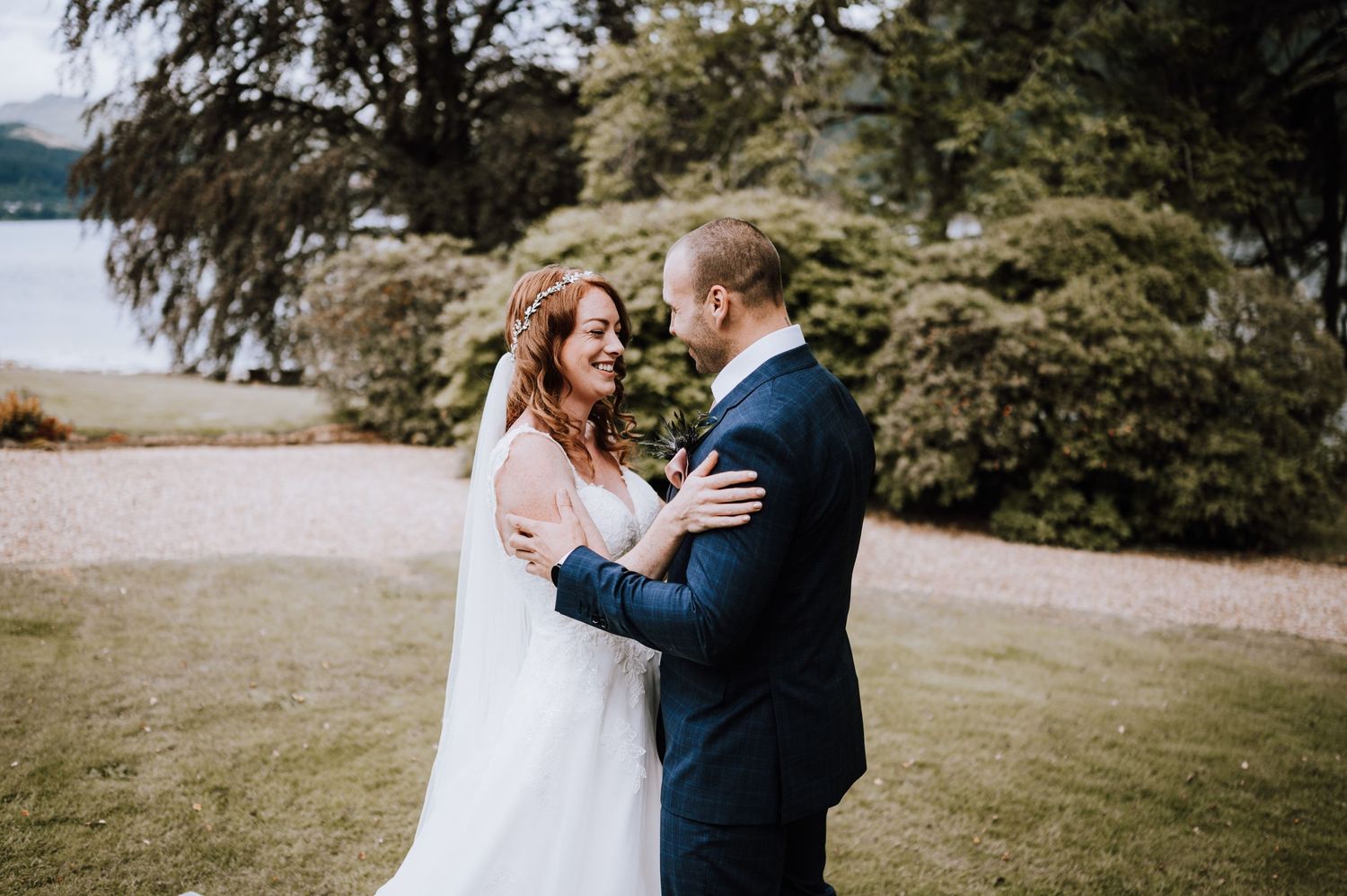 Bride and groom embrace on grassy lawn with trees and water in background.