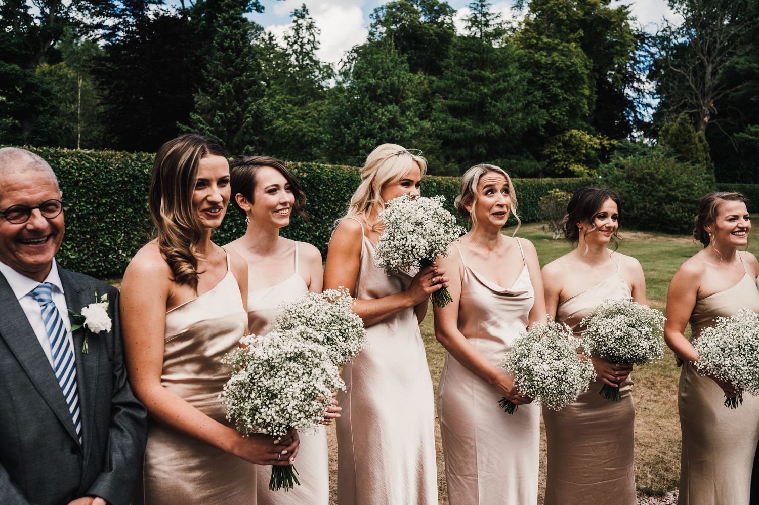 Candid moment of wedding party wearing champagne dresses and holding baby's breath bouquets in garden.