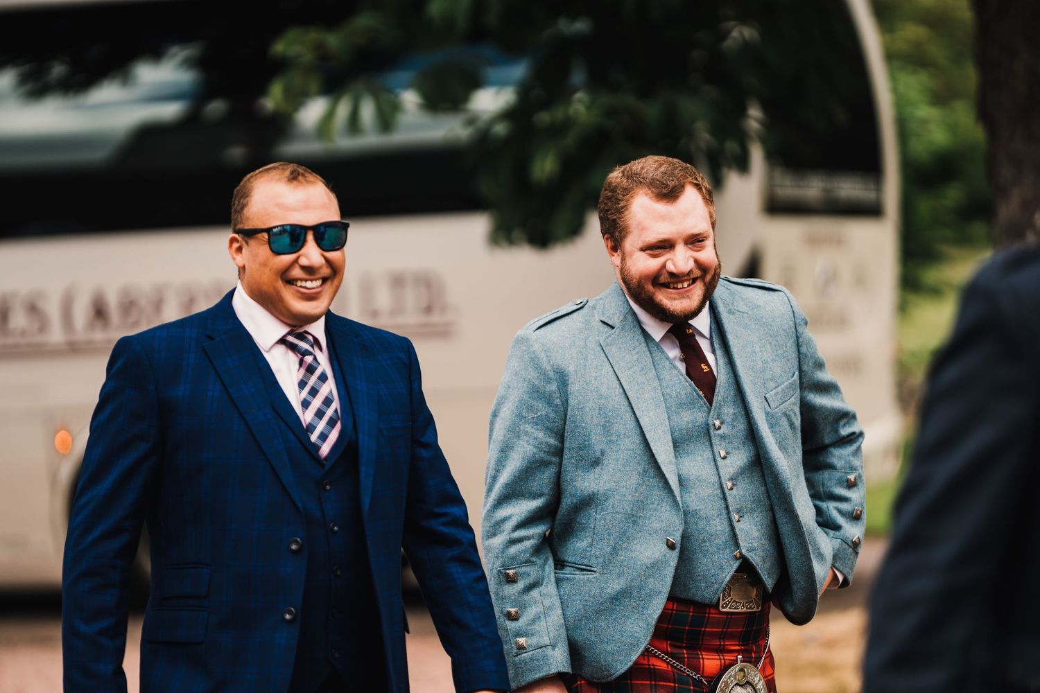 Wedding guests in formal attire and kilts sharing a laugh outside.