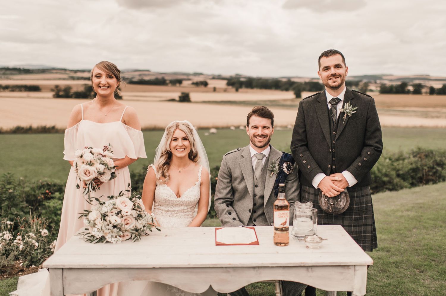 A wedding party stands behind a white table during an outdoor signing ceremony.