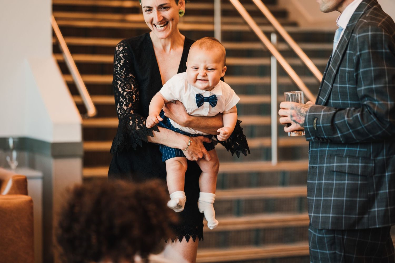 A baby in white clothing smiles during a festive indoor gathering.