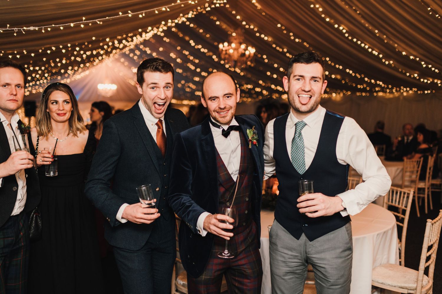 Groomsmen celebrating with drinks at an elegantly lit indoor wedding reception.