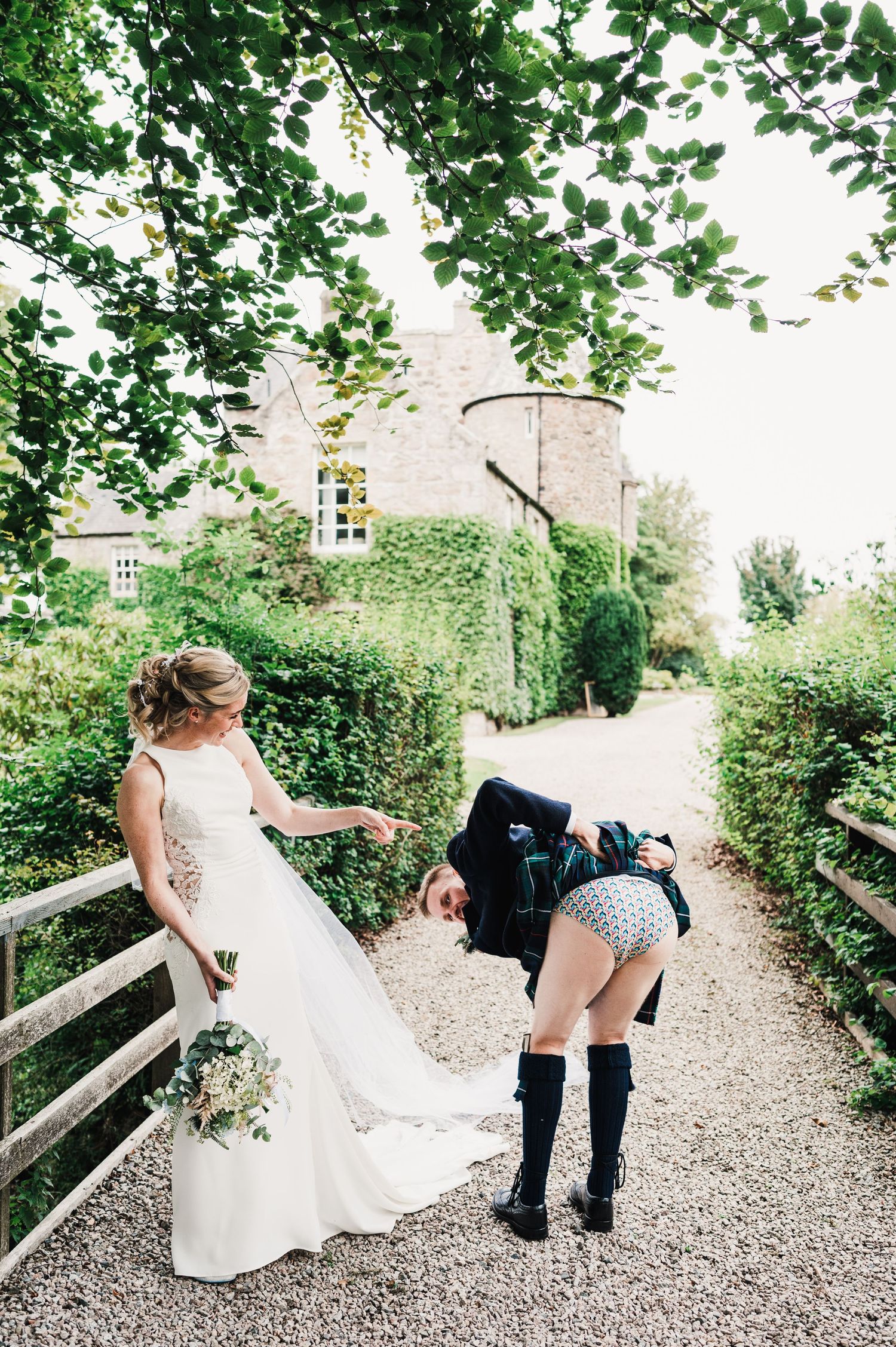 Playful wedding moment on garden path with traditional Scottish attire.