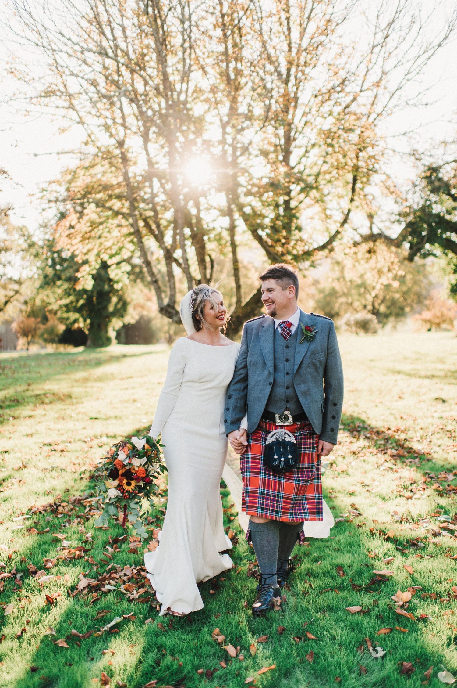 Newlyweds laugh together in autumn sunlight, with the groom wearing traditional Scottish kilt and formal jacket.