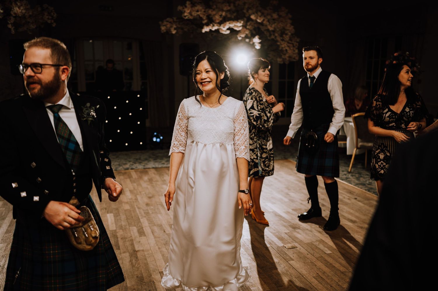 Wedding guests dancing on a wooden floor with twinkling lights in the background.