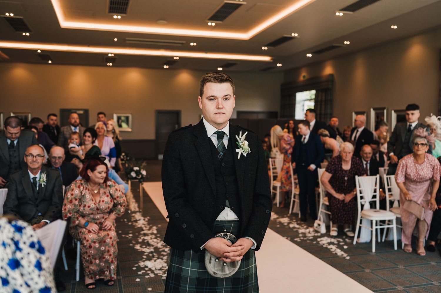 Wedding ceremony in modern venue with Scottish groom in kilt at the altar.