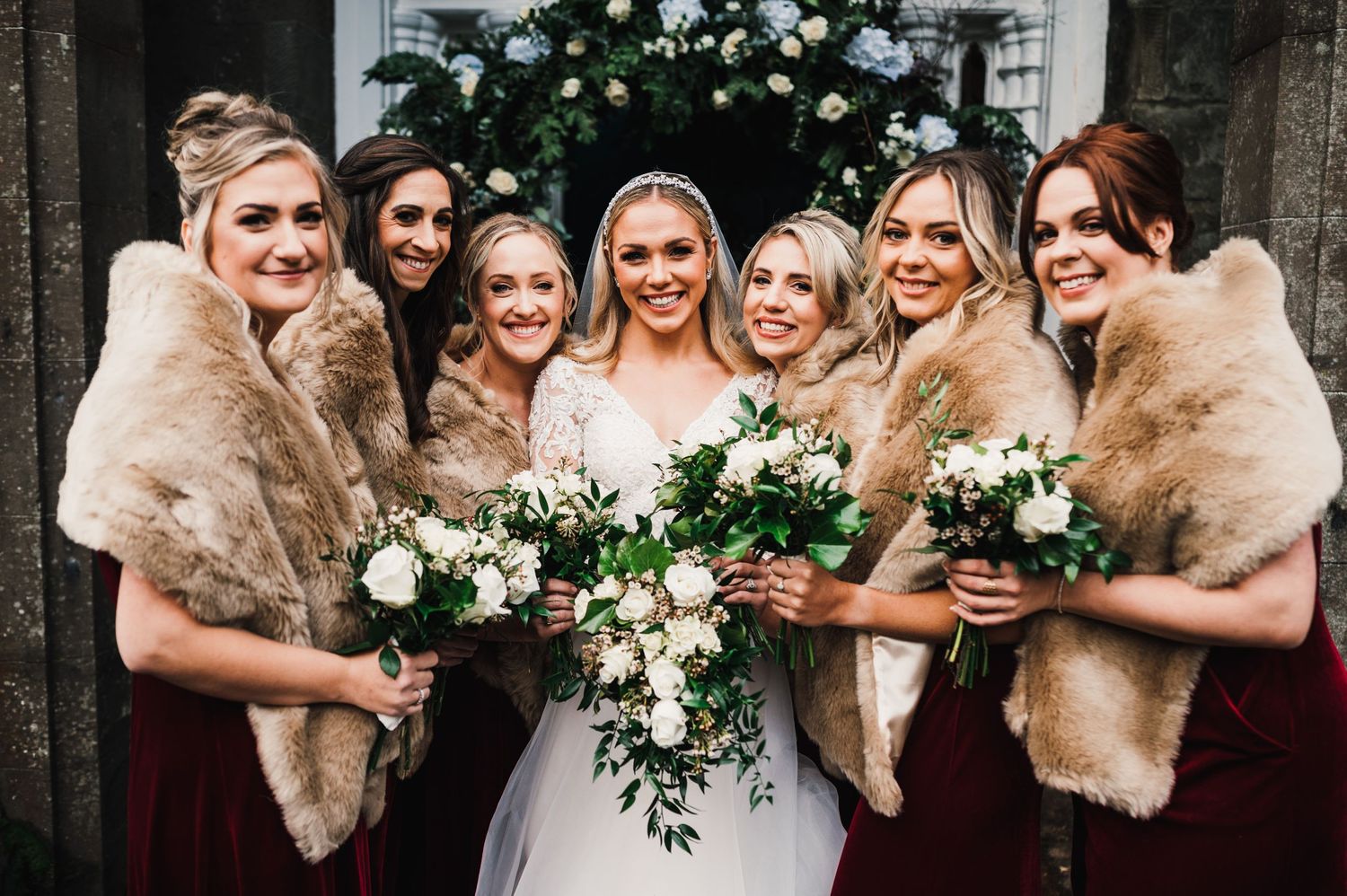Bridesmaids in fur shawls and burgundy dresses surround the bride for a group photo.
