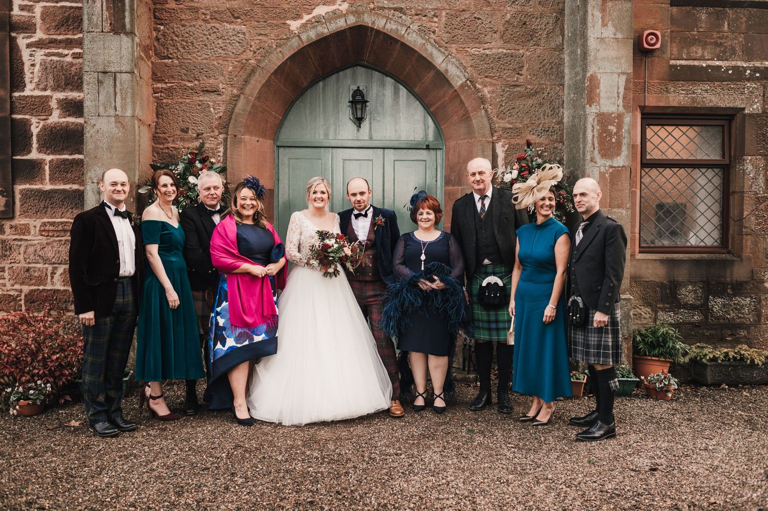 Wedding party in formal attire stands before blue church door with stone archway.