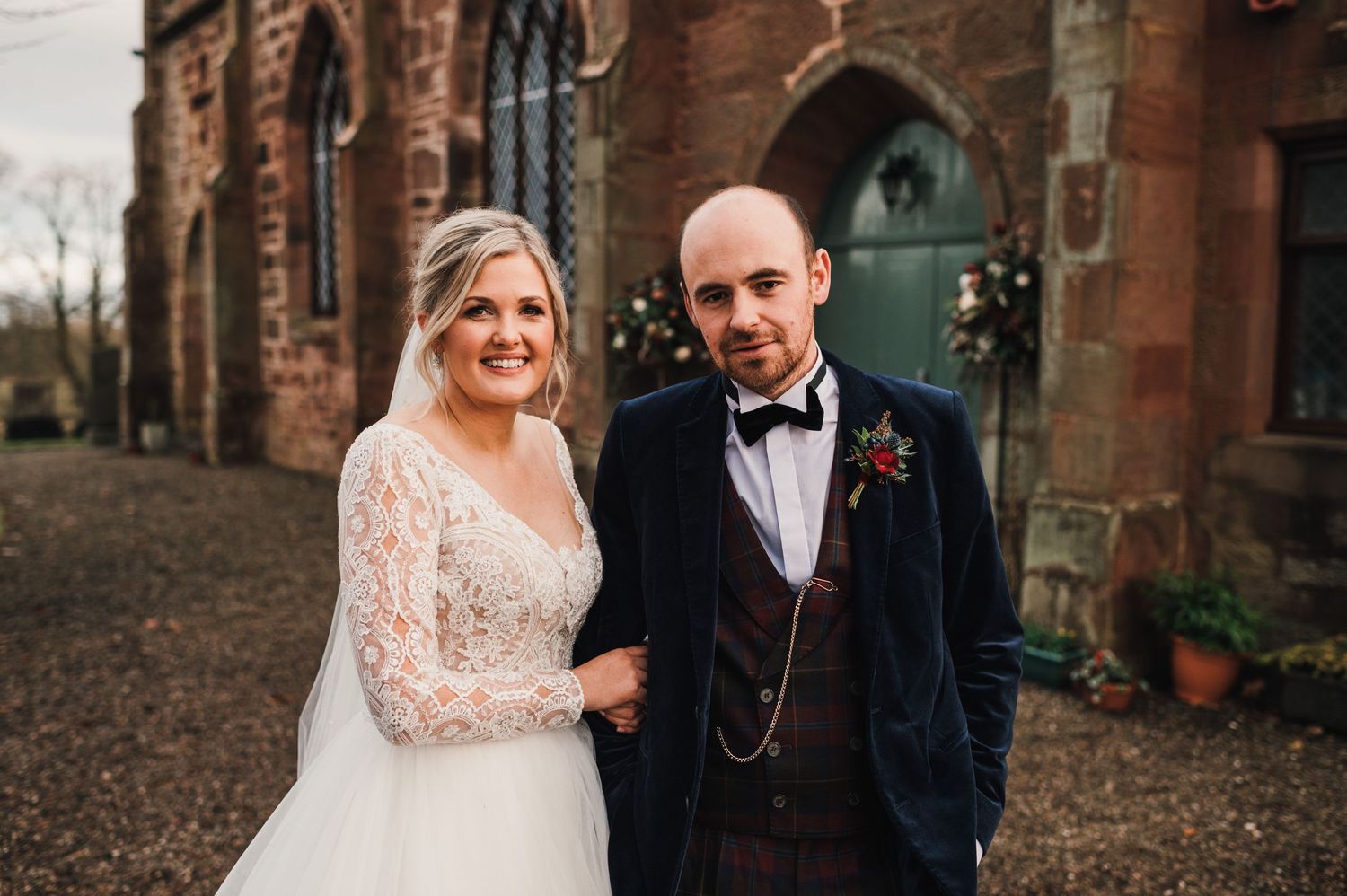 Wedding couple poses in front of rustic church entrance with ornate stone cross detail.