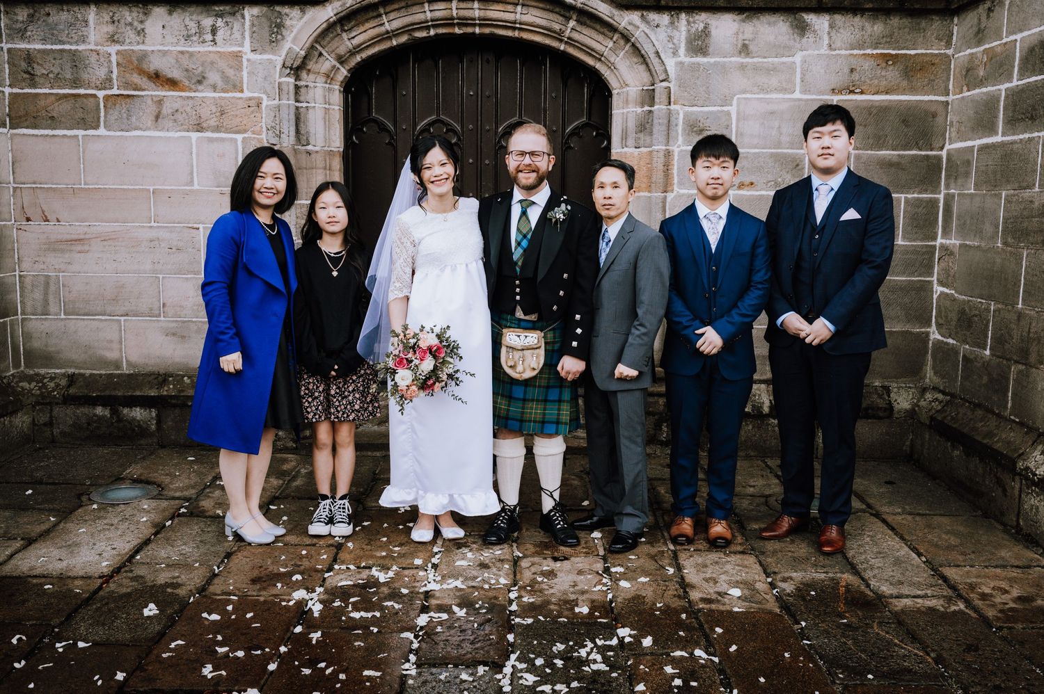 Wedding group photo in front of stone church entrance with groom in traditional Scottish formal wear.