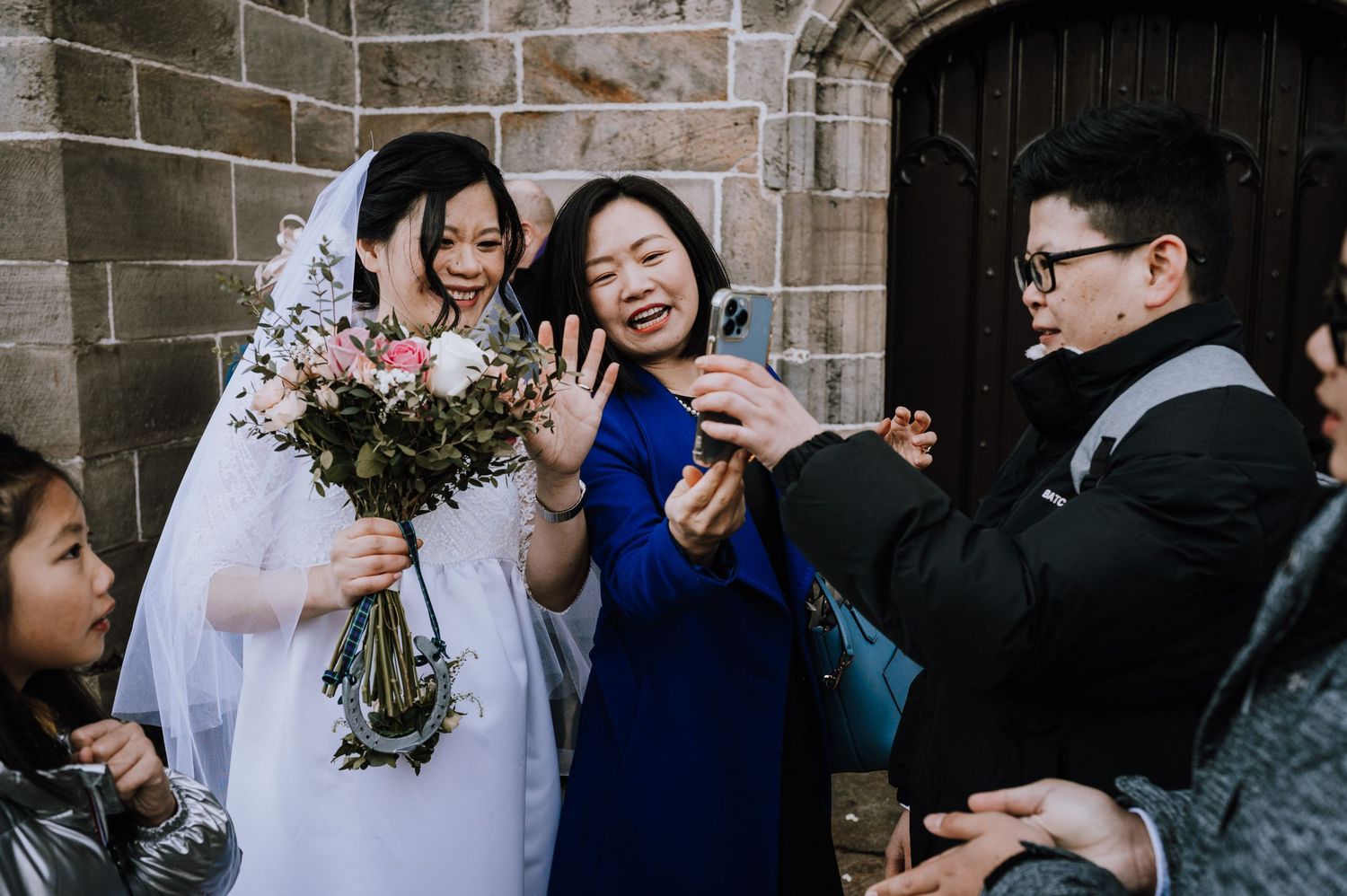 A wedding party gathers outside a stone church taking photos with smartphones.