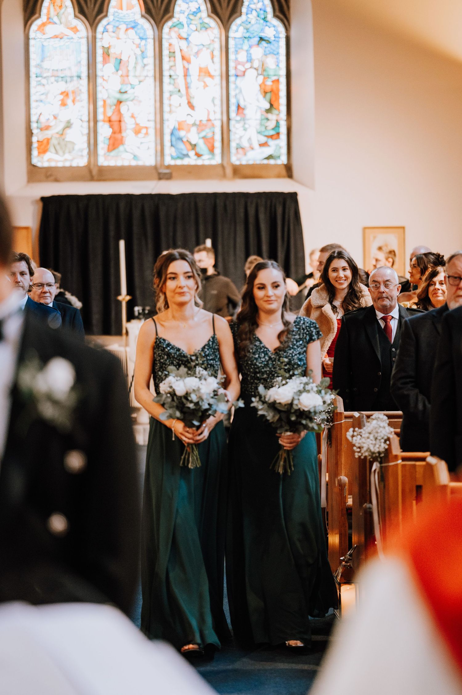 Bridesmaids in elegant emerald green dresses stand at the altar beneath stained glass windows during a church ceremony.
