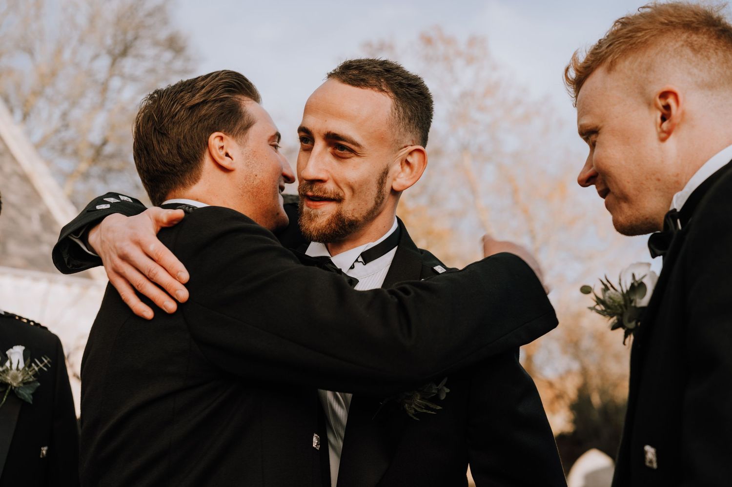 Groomsmen share an emotional embrace outdoors wearing formal black tuxedos.
