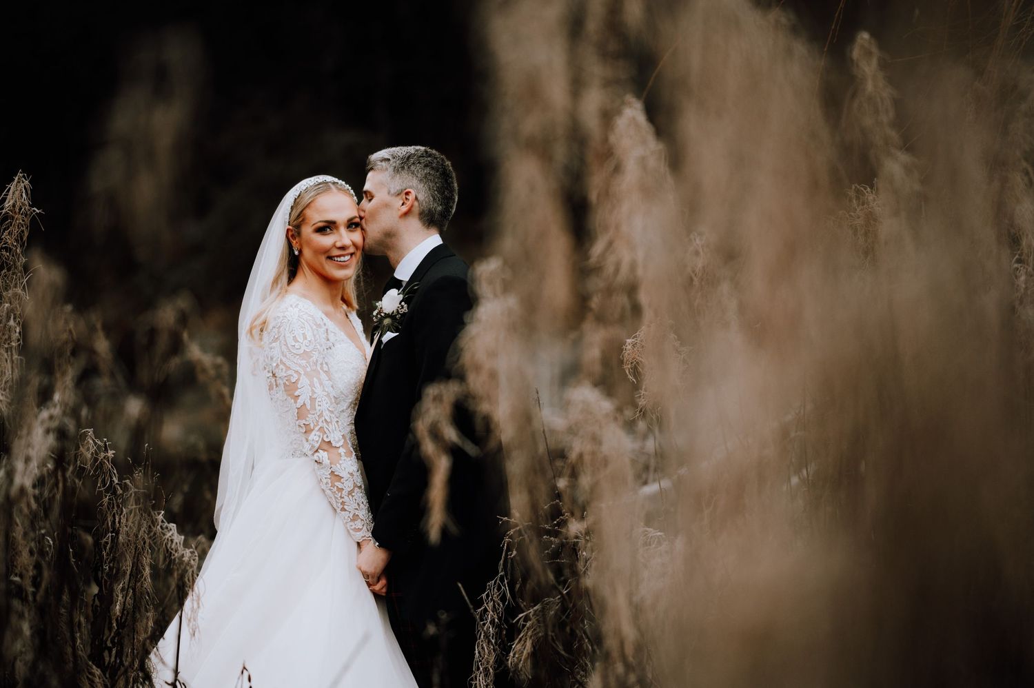 A romantic wedding moment captured among tall dried grasses in natural light.