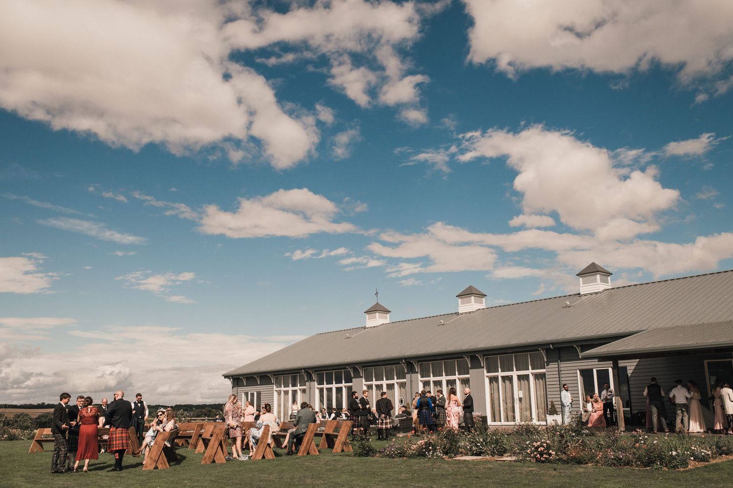 Large barn venue with guests gathered outside under blue skies and white clouds.