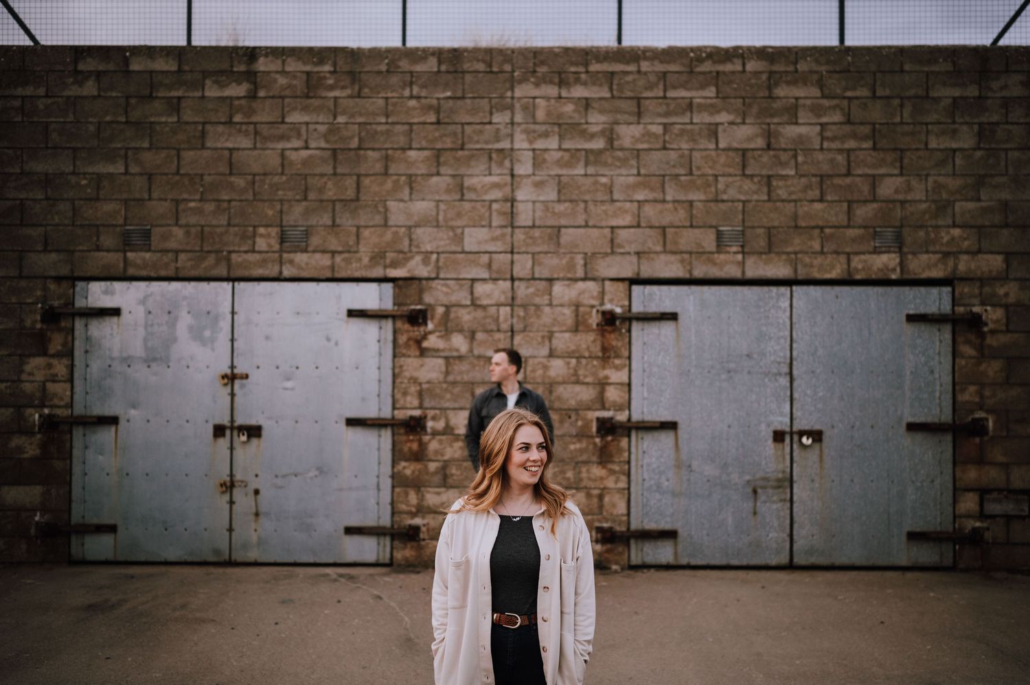 Person in light colored clothing poses against industrial metal doors on brick wall.