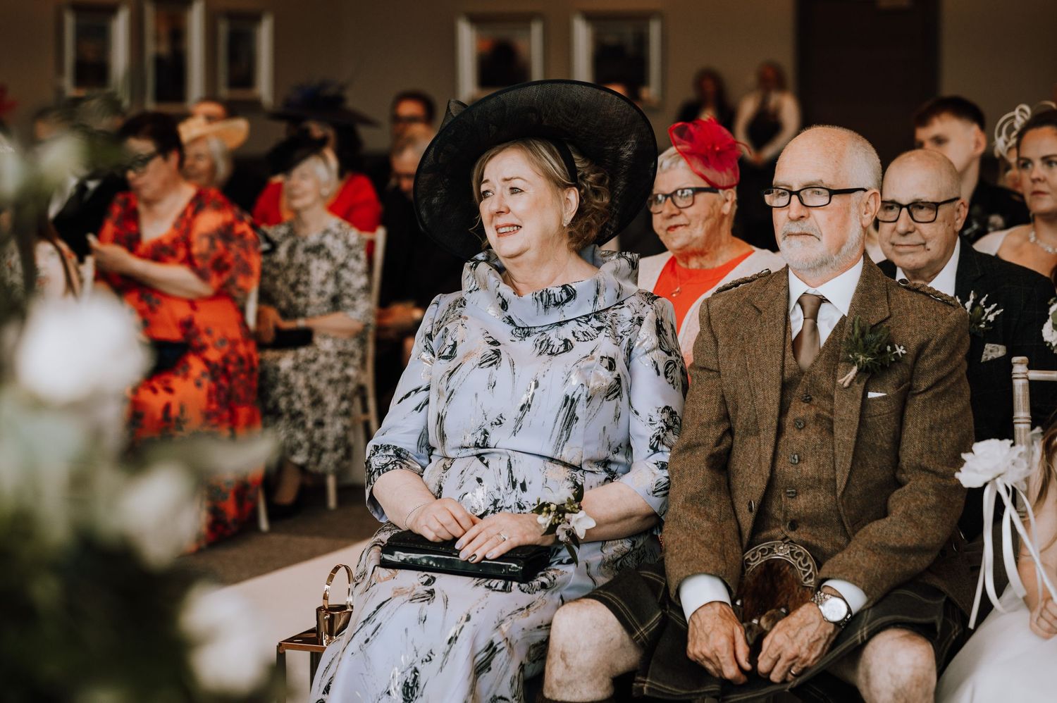 Wedding guests in elegant attire watch the ceremony from decorated chairs indoors.