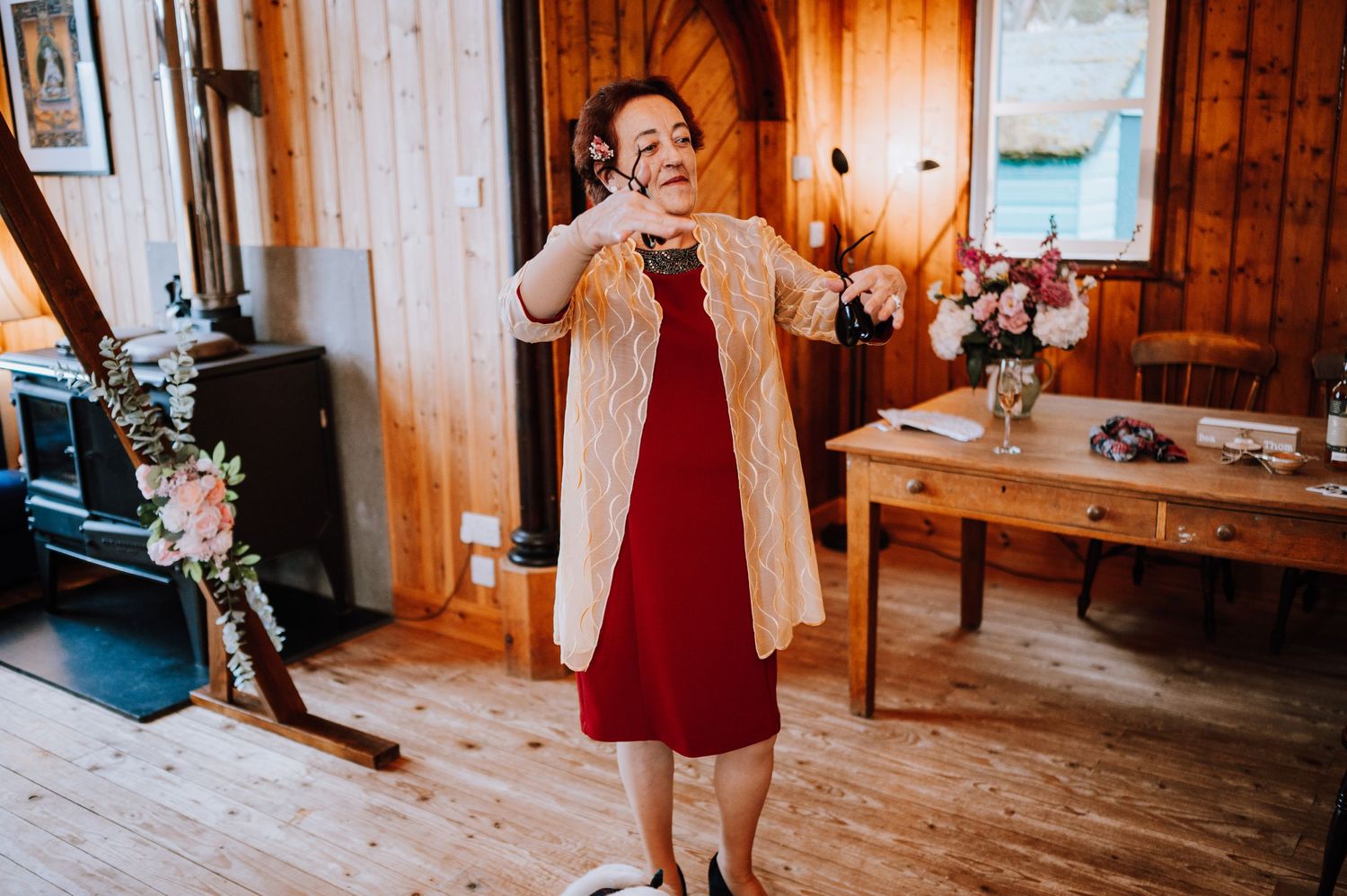 Woman dancing joyfully in a burgundy dress and tan cardigan on wooden floors.