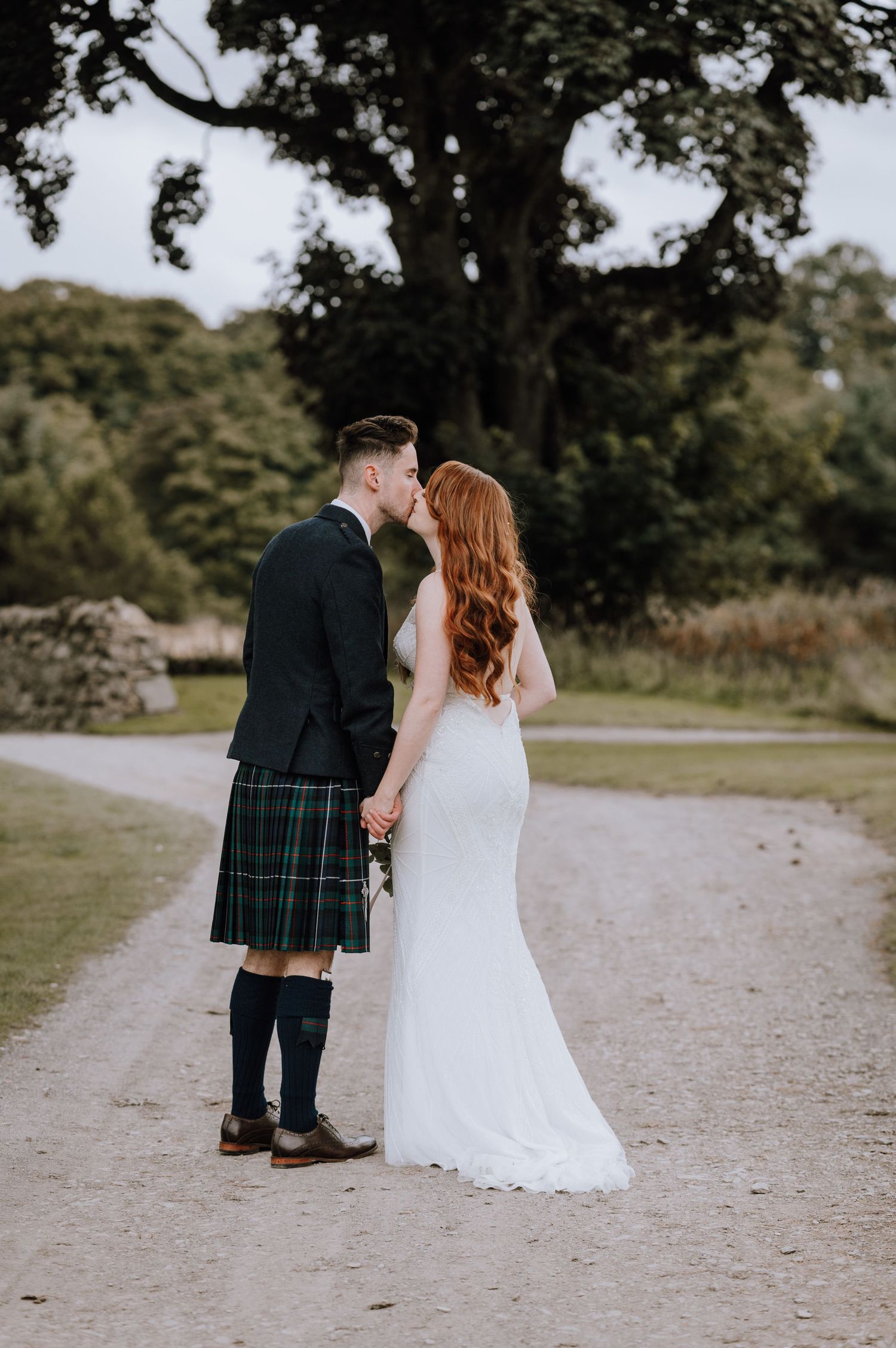 Romantic couple in Scottish wedding attire share intimate moment on castle grounds path.