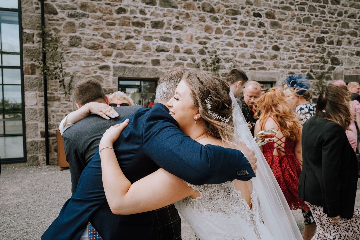 Wedding guests embrace and celebrate during outdoor reception by stone wall.
