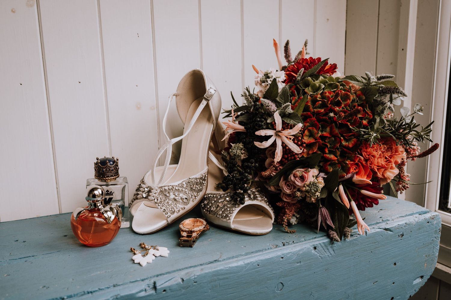 Elegant bridal shoes and bouquet with red flowers displayed on a blue wooden surface.