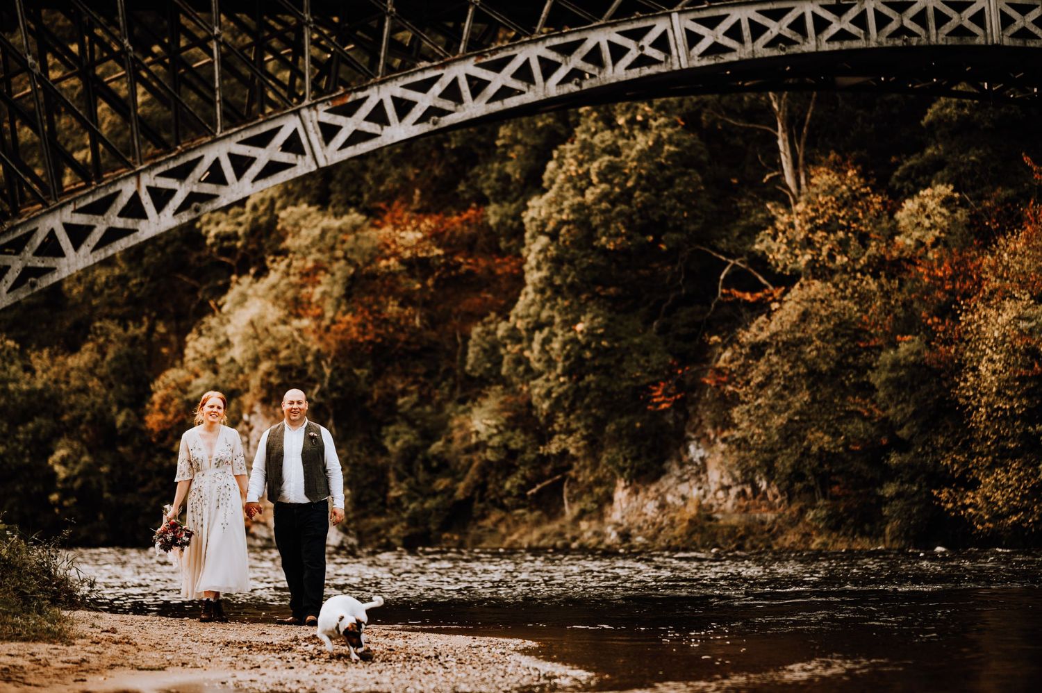 A couple walks with their small dog along a riverside path beneath an iron bridge.