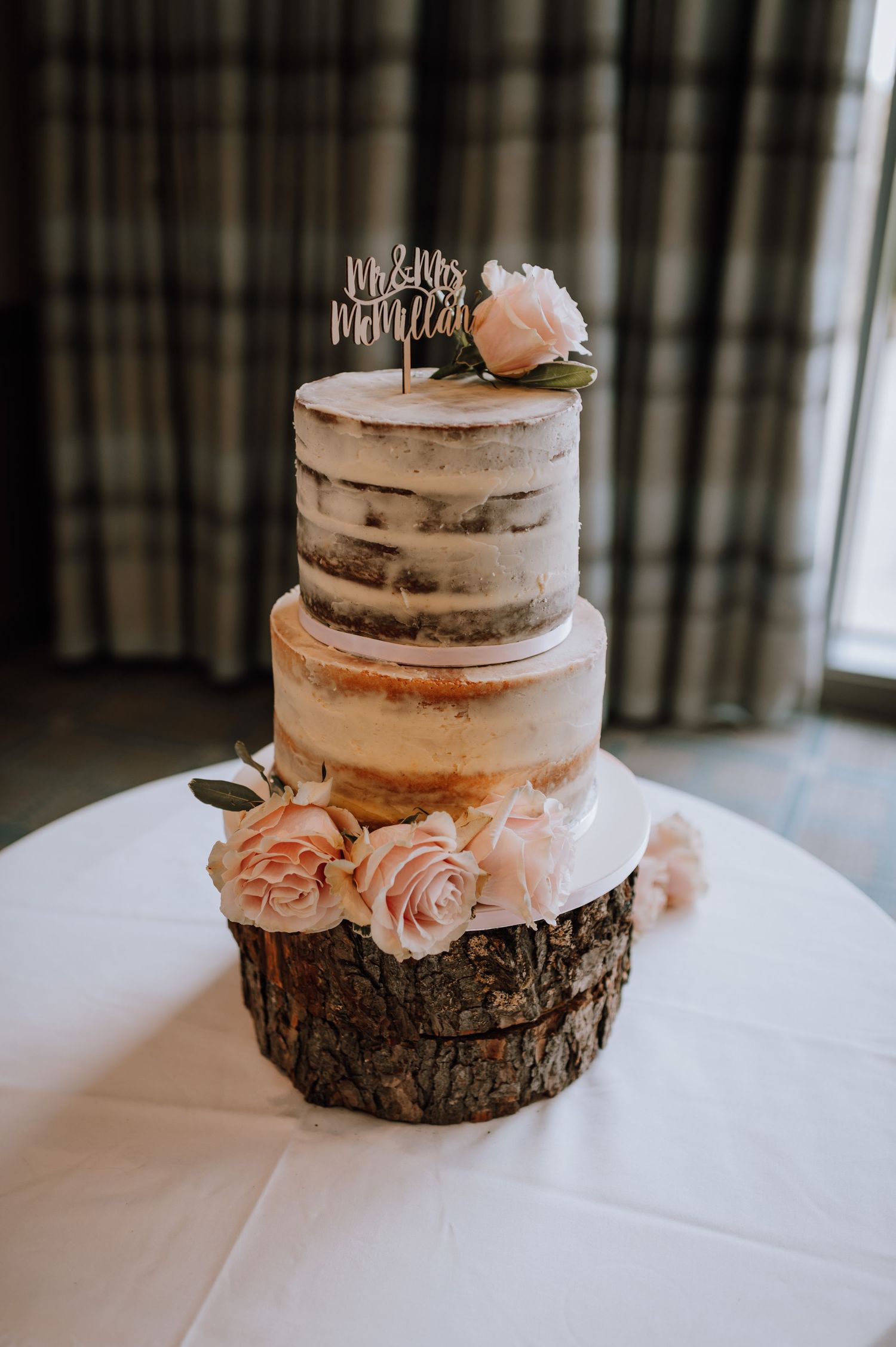 Two-tiered naked wedding cake adorned with pink roses and a cake topper sits on a wooden slice base.