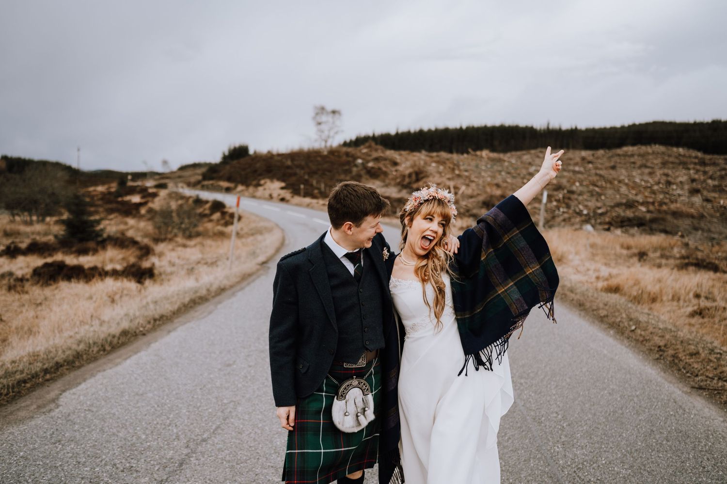 Newlyweds celebrate on rural Scottish road with traditional highland dress.
