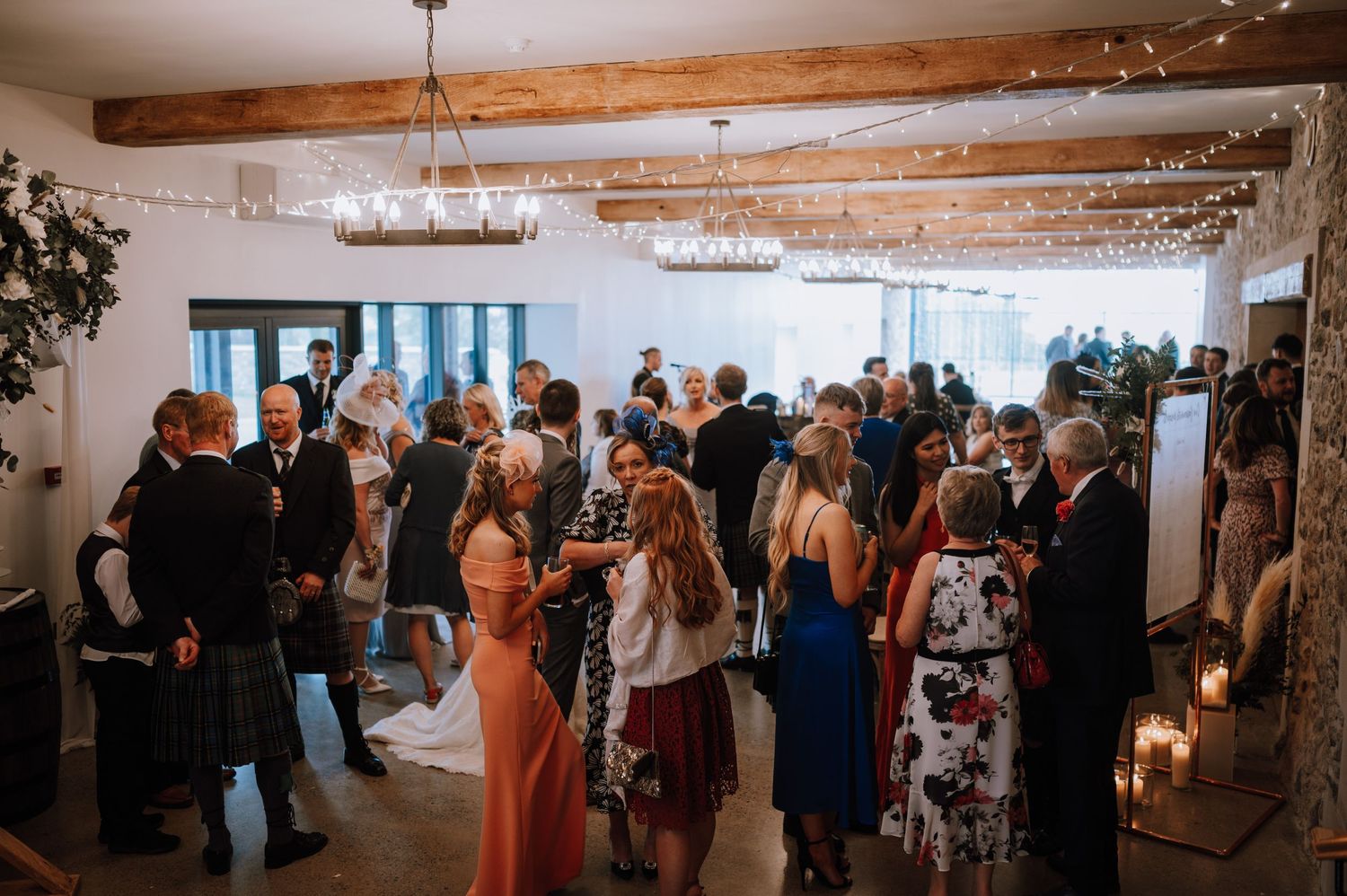 Wedding reception venue filled with guests mingling under exposed wooden beams and chandeliers.