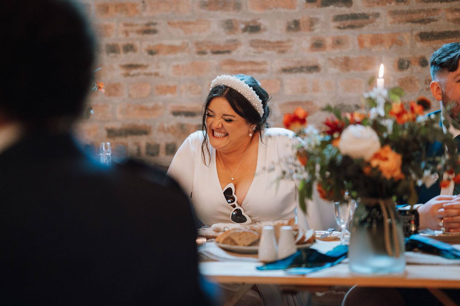 Wedding reception guests enjoy dinner at rustic brick venue with floral centerpieces and blue table settings.
