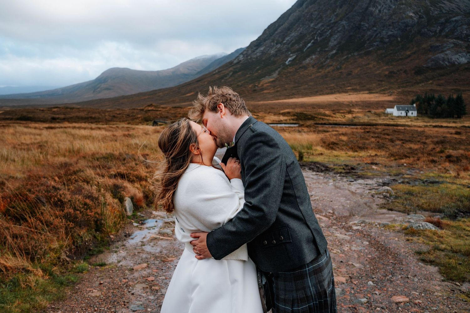 A couple embraces on a scenic mountain path with rugged Scottish Highlands landscapes in the background.