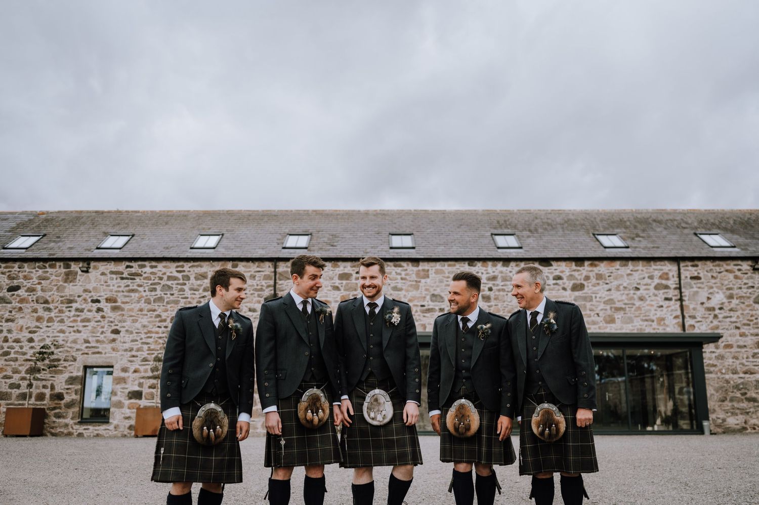 Wedding party in traditional Scottish kilts poses against stone building on overcast day.
