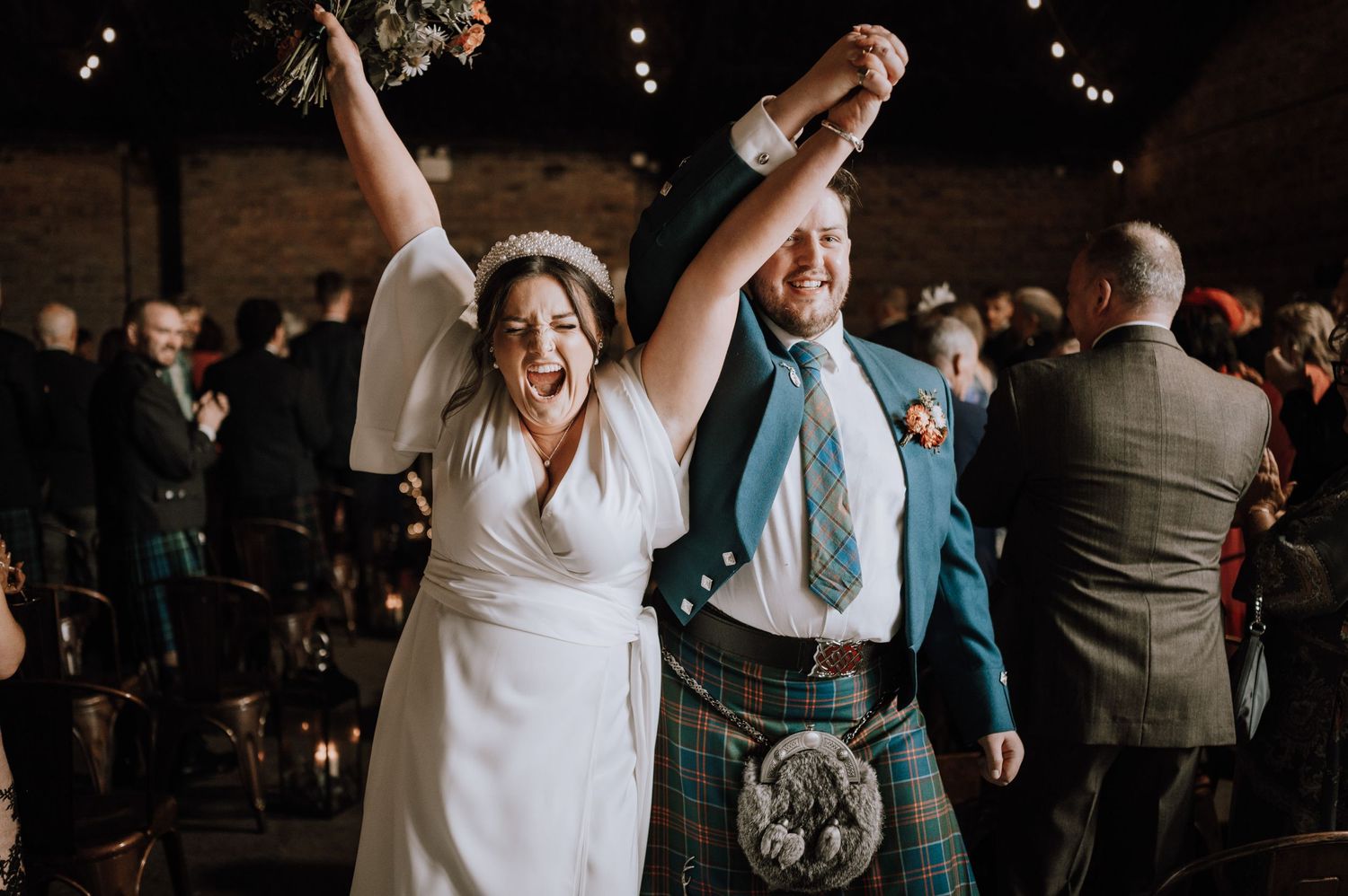 A joyful celebration moment at a wedding with traditional Scottish attire and raised arms.