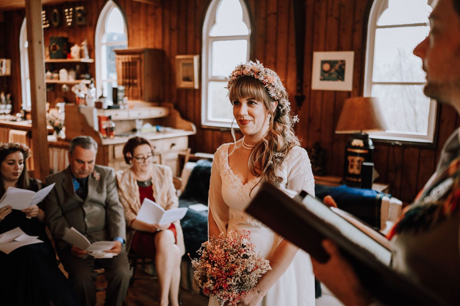A bride in white dress holds flowers during an intimate indoor ceremony.