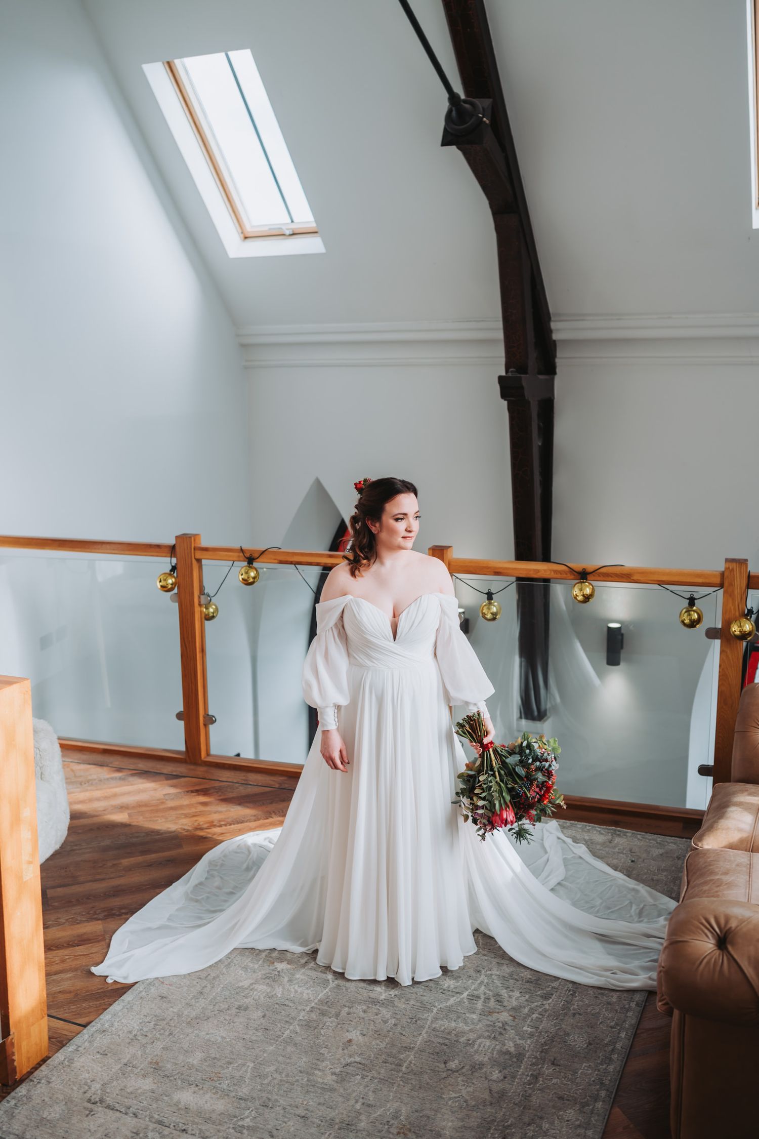 A bride in a flowing white dress stands in a bright attic room with wooden beams and ballet barre along the wall.