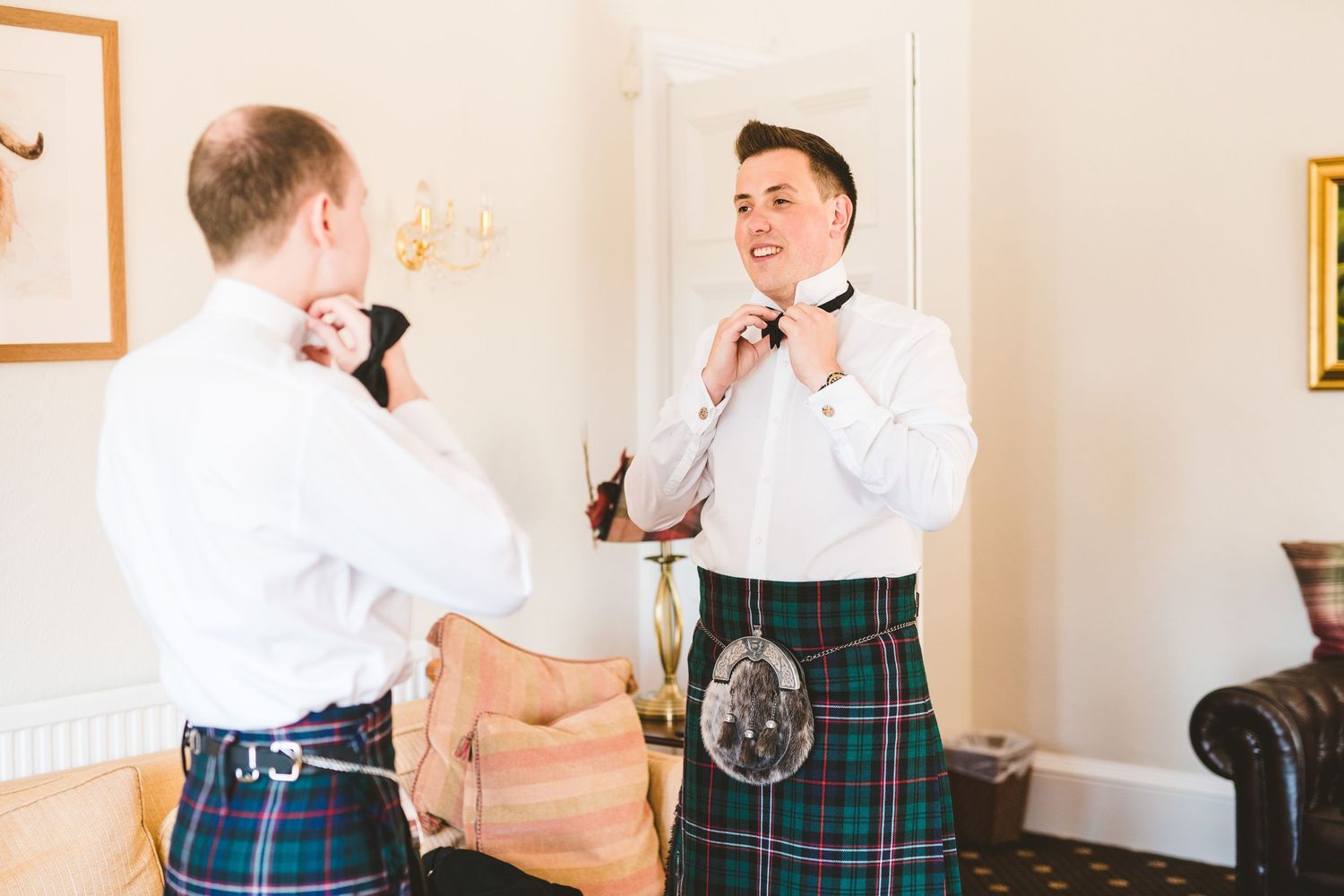 Groomsmen in traditional Scottish kilts help each other prepare and adjust their formal attire before a wedding ceremony.