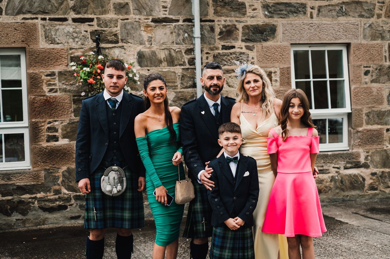 Wedding party in colorful attire poses outside stone building with traditional Scottish elements.