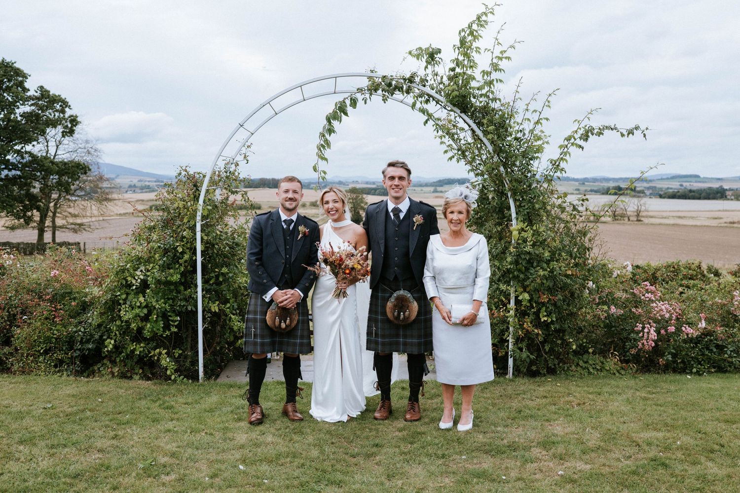 Wedding party poses under floral arch with Scottish highlands in background.