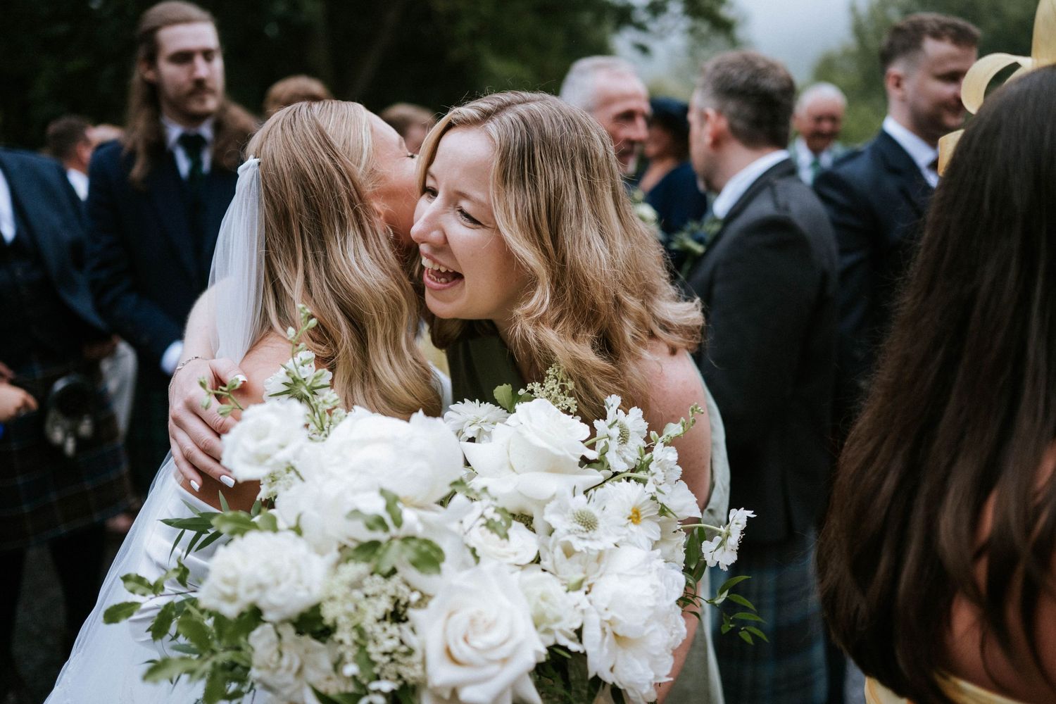 Wedding party shares laughs and embraces while holding elegant white rose bouquets outdoors.