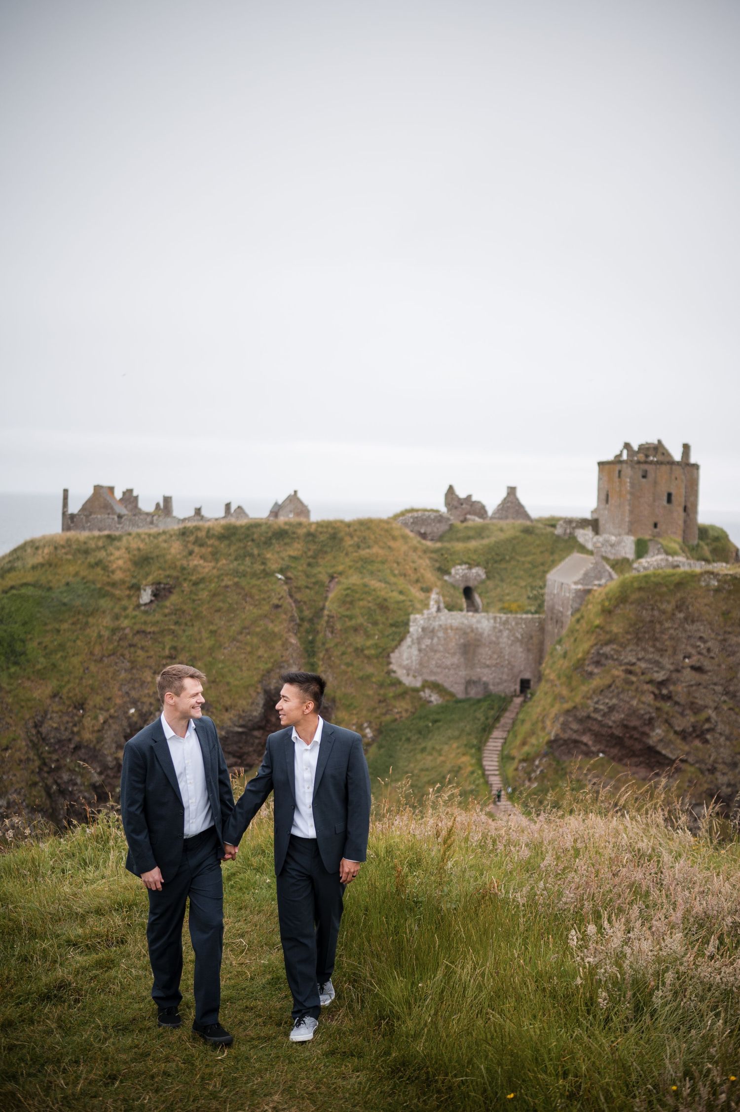 Same Sex Wedding Photography. A couple walks through wild coastal grass with ancient Scottish castle ruins visible on the dramatic cliffs.