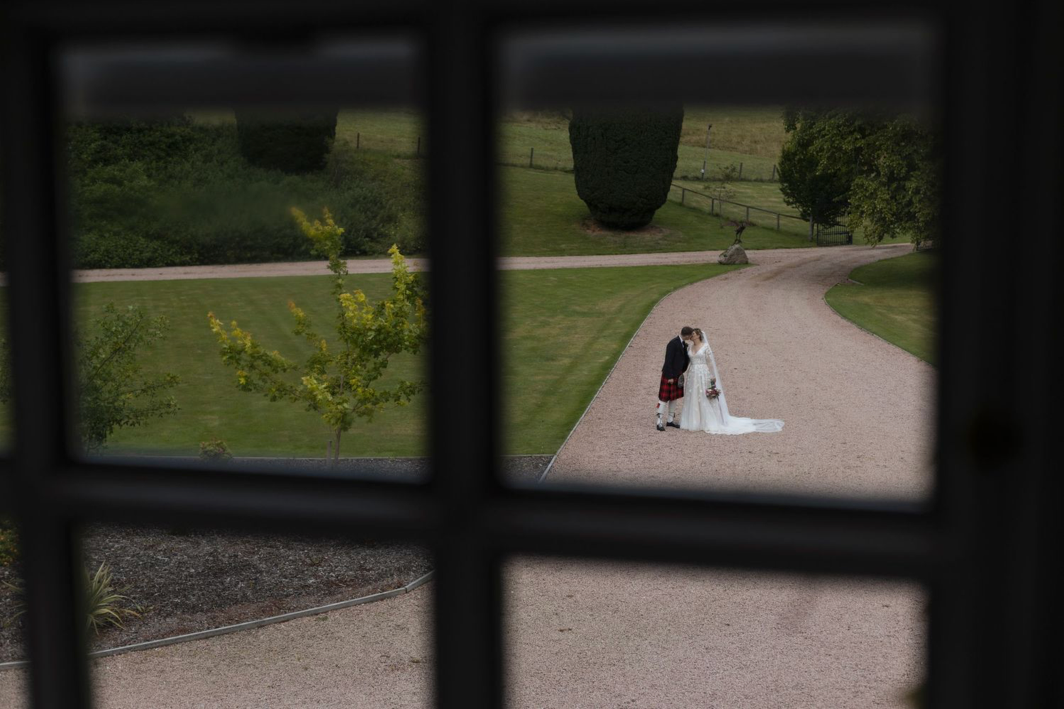 A couple in formal wedding attire walks down a garden path surrounded by lush greenery and trees.