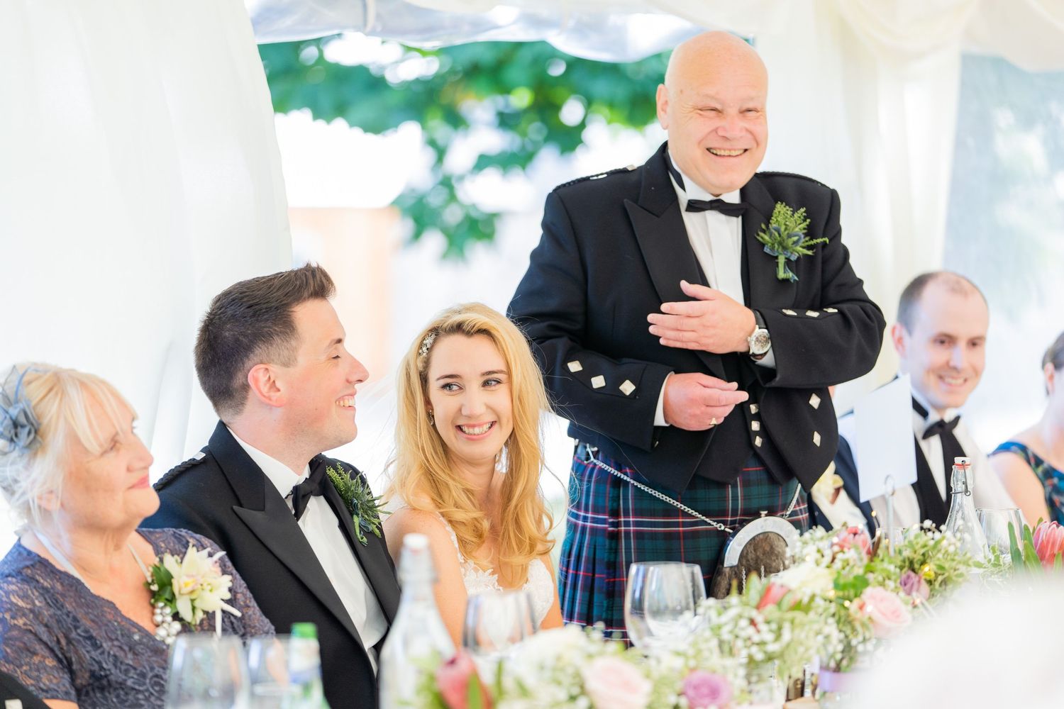 Wedding reception guests seated at tables in a bright white marquee tent.